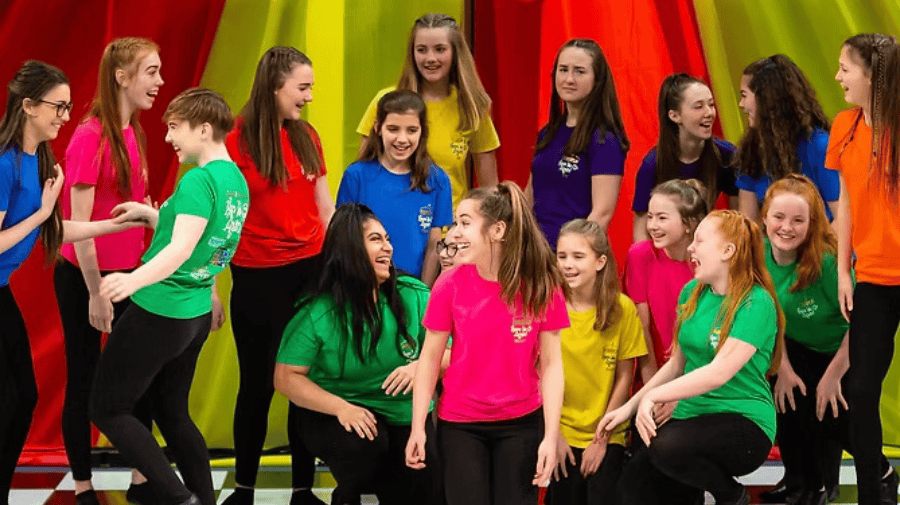 a group of teens in bright tops talk animatedly to each other against a circus tent background