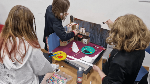 three children with their backs to the camera set up a stop-motion animation set, on a Halloween theme