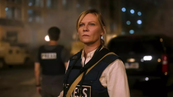 a blonde middle aged woman stands in a city street at night. She's wearing a flack jacket with PRESS on it and is staring off into the distance 