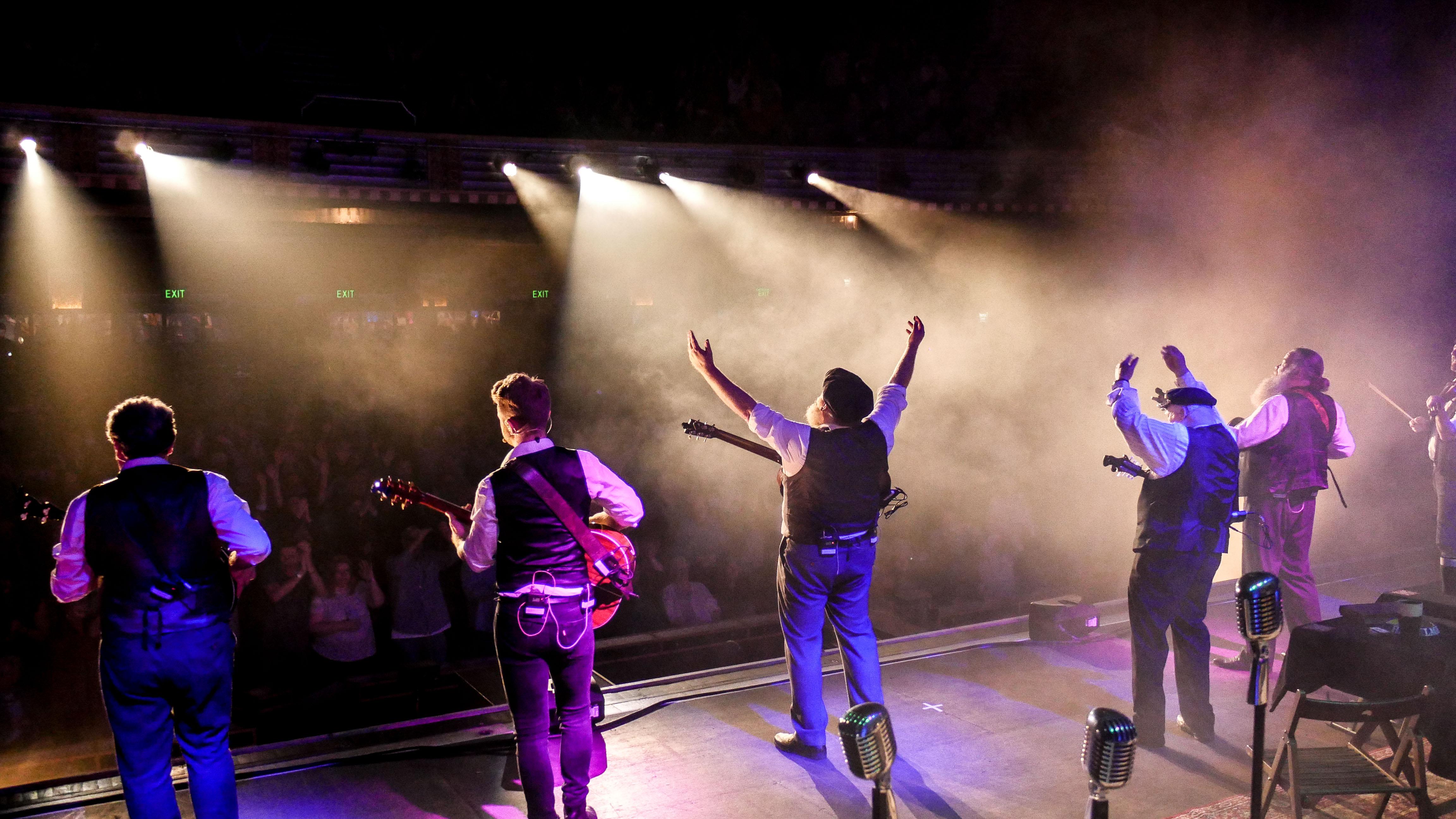 5 people stood on stage playing instruments or arms in the air