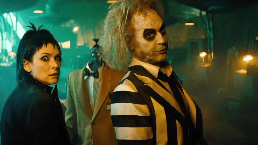 winona ryder, in character, all in black looks over her shoulder to the camera. Michael Keaton as Beetlejuice is in a white suit with thick black stripes and has wild green hair and a painted white face 