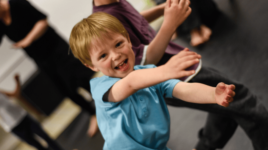 a young child with short blonde hair and a blue top smiles enthusiastically and throws their arms out 