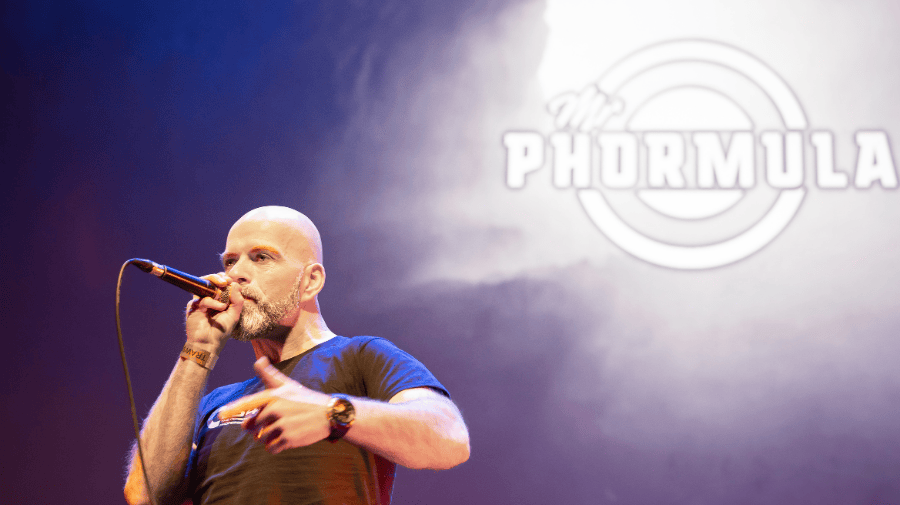 Mr Phormula, shaved head wearing a dark t-shirt, is holding a microphone to his mouth. Behind, a projection of his stage name in white light 