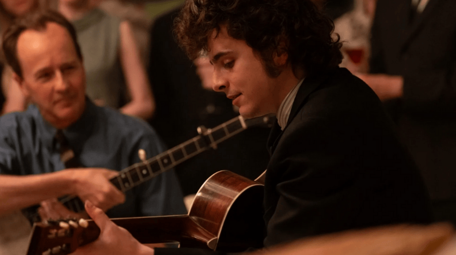 a young man with dark curly hair smiles as he plays guitar. He's facing side on, wearing a dark jacket. He's watched intently by another man 