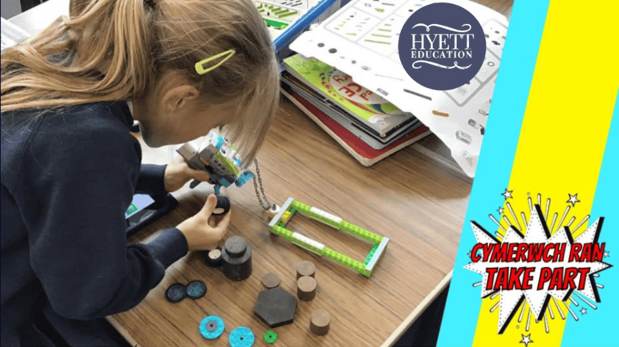 a young girl with a blonde ponytail, in school uniform jumper, bends over a table working on Lego and electronics 