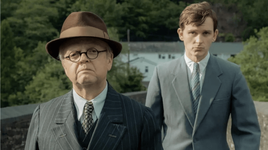 Toby Jones as Mr Burton, a round-faced white man, wearing a 1940s sports jacket, brown trilby and shirt, tie and jumper. Behind him a young white man with brown hair in similar clothes. they are stood on a pathway with green trees around them