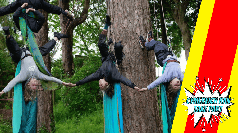 a group of young people with long hair hang upside down from ropes against large tree trunks with turqouise drapes around them 