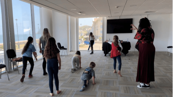 a room with large floor to ceiling windows and two adults with a group of young children running about backs to the camera