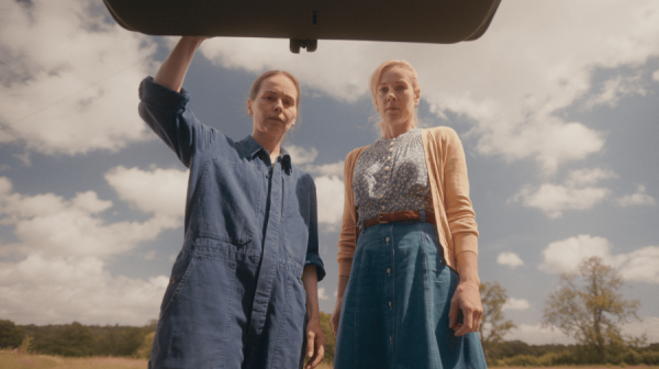 two women hold open the boot of a vehicle and look in, both very serious.One is wearing blue overalls, the other is in skirt, blouse and cardigan. Behind them, an open field