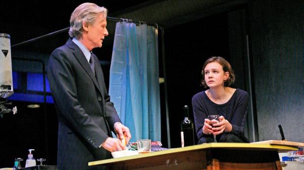 Bill Nighy, in a dark suit, stands at a table. Carey Mulligan, in a black top, leans on the same table. Behind them is bath with a plastic curtain pulled around it.  