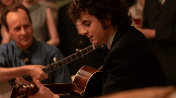 a young man with dark curly hair smiles as he plays guitar. He's facing side on, wearing a dark jacket. He's watched intently by another man 