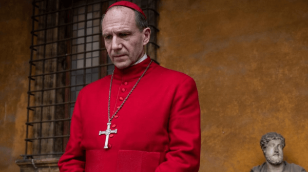 Ralph Fiennes, a white male actor in his 60s, is dressed in a Bishop's surplice. It is bright red. Around his neck is a Cruxifix. Behind him, a wall painted ochre yellow 