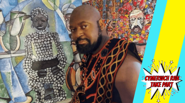 a black man with a shaved head wearing traditional african clothes stands in front of colourful African artwork 