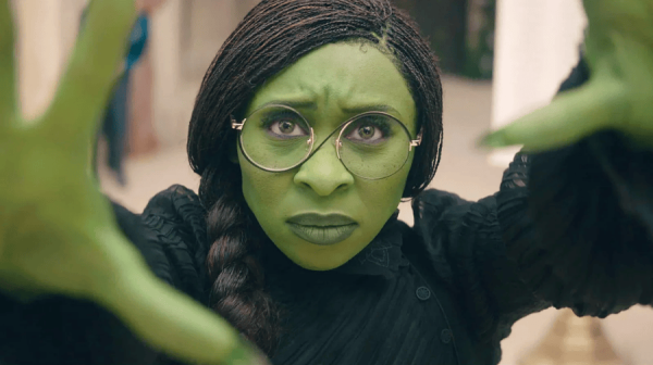 a young woman with green skin holds her hands up to the camera. She has wire rimmed glasses on and braided black hair 